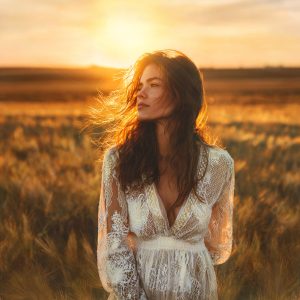 Golden hour feminine artwork of a woman in a white lace dress standing in a sunlit field, symbolizing stillness, inner peace, reflection, and quiet transformation.